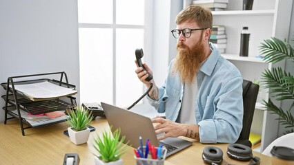 Wall Mural - Gritty irish boss, adult redhead man, elegantly talking on the phone while working on laptop at office desk, indoor setting