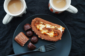 Chocolate roll with cream and caramel next to chocolate on a black plate near two cups of coffee with milk