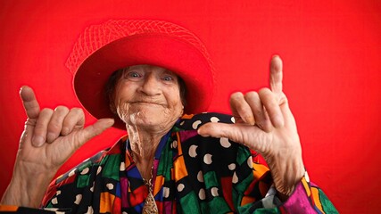 Wall Mural - Fisheye portrait of funny elderly mature woman, 80s, wearing red hat giving shaka hand gesture isolated on red background. Concept of old rock and roll person