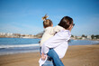 Happy mother giving piggyback ride to her lovely daughter, enjoying happy family time together on the Atlantic beach.