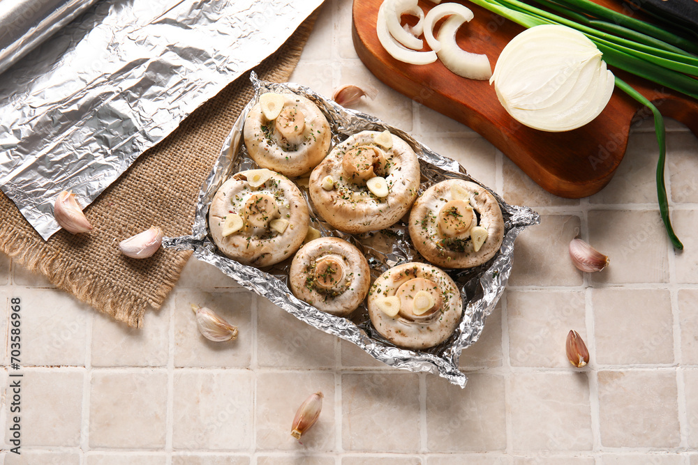 Aluminium foil with raw mushrooms, onion and garlic on light tile background