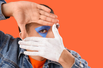 Wall Mural - Young man with painted body making frame gesture on orange background, closeup