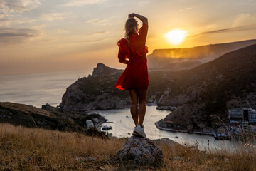 Wall Mural - Happy woman standing with her back on the sunset in nature in summer with open hands posing with mountains on sunset, silhouette. Woman in the mountains red dress, eco friendly, summer landscape 