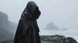  a person in a black hooded cloak standing on a rocky beach with a body of water and mountains in the background.