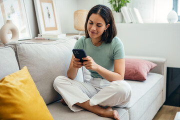 Wall Mural - Smiling young woman using her mobile phone while sitting on sofa at home