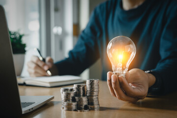 Wall Mural - Businessman holding and putting lightbulb on coins stack on table for saving energy and saving money concept.