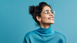 Horizontal photograph of a self embracing and joyful woman who appears optimistic but shy donning transparent glasses and a casual turtleneck standing alone against a blue background 