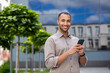 © Liubomir - Portrait of young businessman, man smiling looking at camera holding phone, worker using app on smartphone while walking in city close up outside office building.