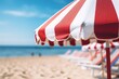 Close up image of a red and white vertical striped beach umbrella on a background of a beach with sun loungers and blue sea