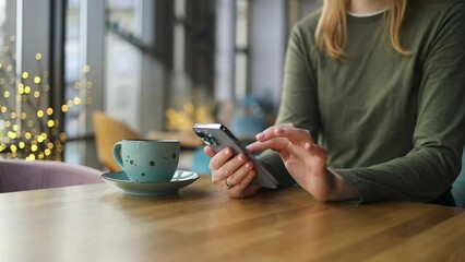 Crop view of female person holding smartphone and scrolling mobile phone while sitting at table with coffee at coffee shop cafe. Work people contact marketing business, technology. High quality FullHD