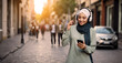 © ginettigino - Joyful young muslim woman in hijab listening to music through headphones connected to cell phone while walking around the city center.