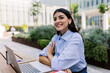 © Xavier Lorenzo - Smiling woman looking away while working online with laptop sitting outside of his office building. Business and entrepreneur people concept.
