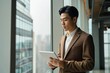 Young Japanese business man, Asian company manager or entrepreneur wearing suit holding digital tablet pc standing near big office window checking financial services or corporate, Generative AI 