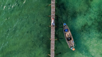 Wall Mural - Aerial view of a person lying on a wooden pier next to a small boat in clear turquoise waters, conveying a sense of relaxation and solitude