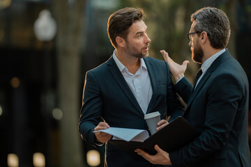 Wall Mural - Photo of business partners talk conversation outdoor. Two handsome businessmen in suits discuss a working project against the background of urban city. New projects or startups. Cooperation concept.