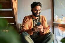 Happy Indian Ehnic Man Relaxing At Home Drinking Coffee Wearing Earbud Using Cell Phone Having Virtual Mobile Chat Video Call On Smartphone, Watching Social Media Videos Or Game, Generative AI 