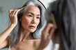 Senior attractive middle 50 years aged asian woman with gray hair looking at mirror reflection combing tangled gray hair. Alopecia hair loss prevention treatment after menopause, Generative AI