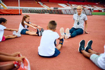 Wall Mural - Happy sports teacher and group of children doing relaxation exercises at stadium.