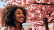Modern happy young smiling dark-skinned African woman with a glass of coffee against the backdrop of pink cherry blossoms and metropolis city.