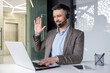 © Liubomir - A young man sits in the office at a desk in a headset and works on a laptop, talking and greeting to the camera online.