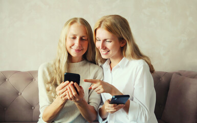 Two caucasian women with mobile phone looking at device, middle aged mother or sister and adult daughter together sitting on couch in living room at home