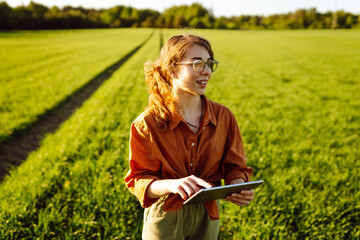 Wall Mural - Smart agriculture. A young woman farmer wearing glasses holds a tablet on a green wheat field. A woman agronomist checks the quality of the crop using a digital tablet. Agriculture concept.