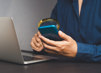 Wall Mural - businessman holding a magnifier and looking at a smartphone while sitting at a desk.