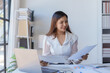 © amnaj - Smiling asian business woman accountant analyst holding documents, work at laptop computer doing online trade market tech research.
