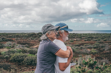 Canvas Print - Bonding senior couple in walking day in countryside looking at the horizon over water. Elderly smiling retirees enjoying healthy lifestyle and vacation in nature