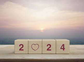 Wall Mural - Love icon with 2024 letter on wood block cubes on wooden table over city tower and skyscraper at sunset, vintage style, Happy new year 2024 and Valentines day concept