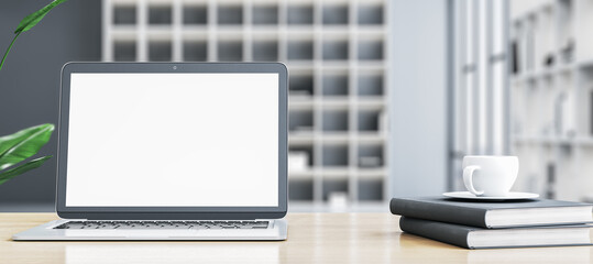Poster - Modern wide designer office desktop with empty white mock up laptop screen, coffee cup on stacked books and other items on blurry office background. 3D Rendering.