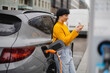 © Westend61 - Woman using smart phone and leaning on car at electric vehicle charging station