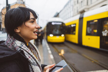 Happy Woman Holding Smart Phone And Waiting At Tram Station