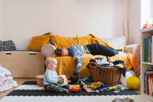 Exhausted Mother Sleeping On Sofa With Baby Boy Playing At Home