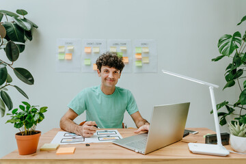 Smiling graphic designer sitting with laptop and printout at desk in office