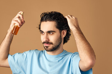 Hair repair treatment. Serious young man applying essential oil spray on his curly brown hair against beige studio background. Concept of male beauty care, hair care cosmetic product, hygiene.