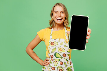 Wall Mural - Young smiling housewife housekeeper chef cook baker woman wear apron yellow t-shirt hold use blank screen mobile cell phone blink eye isolated on plain pastel green background. Cooking food concept.