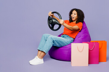 Full body little kid teen girl wears orange t-shirt sit in bag chair hold steering wheel drive car shopping paper package bags isolated on plain purple background. Black Friday sale buy day concept.