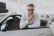Adult man customer male buyer client wearing shirt glasses open lean on door prop up chin choose auto want buy new automobile in car showroom vehicle salon dealership store motor show. Sales concept.