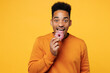Young excited overjoyed man wear orange sweatshirt casual clothes hold in hand eat bite donut isolated on plain yellow background studio. Proper nutrition healthy fast food unhealthy choice concept.