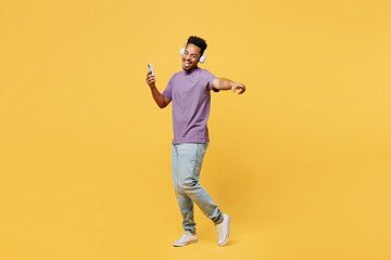 Wall Mural - Full body fun young man of African American ethnicity he wears purple t-shirt casual clothes use mobile cell phone listen to music in headphones isolated on plain yellow background. Lifestyle concept.