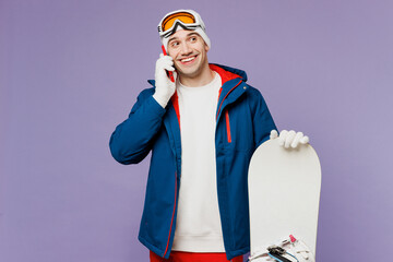 Smiling man wear warm blue windbreaker jacket ski goggles mask hat talk speak on mobile cell phone hold snowboard spend extreme weekend winter season in mountains isolated on plain purple background.