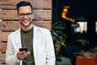 Portrait of young man professional CEO looking at camera and using smartphone while standing in front of restaurant.