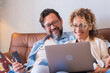 © simona - Portrait of two people man and woman enjoy technolofy connection toether with laptop and phone smiling and watching the computer - happy cheerful couple at home sitting on the sofa at work