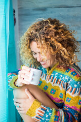 Wall Mural - Young adult woman at home sitting and drink - smile happy caucasian female people enjoy indoor activity with relax and wellbeing alone - blue tones and window light lifestyle portrait