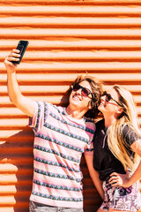 Wall Mural - Young modern cheerful couple man and woman take together a selfie picture in outdoor wirh colorful orange and blue wall in background - beautiful and youthful caucasian people enjoy sharing 