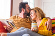 © simona - Happy beautiful caucasian couple at home enjoy morning leisure activity in relax together - love and relationship life with young people sitting on the colorful sofa with book and coffee