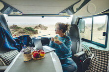 Living And Working Inside Your Camper While Traveling And A Digital Nomad-free Lifestyle. A Woman Sitting In An RV Enjoys Relaxing And Connecting To Her Laptop. The Beach In The Background Outside