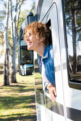 Sticker - Beautiful smiling Caucasian curly woman leaning out of the window of a camper. Travel, nature and leisure concept