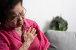 Asian senior elderly grandmother suffering from heart pain and holding her chest while sitting on the sofa in her living room. Medical emergency treatment and health care insurance protection concept